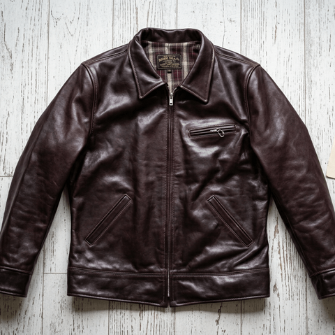 A dark brown leather jacket with a zippered front and chest pocket, laid flat on a white wood plank background next to a small handwritten note.