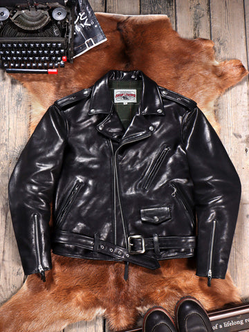 A black leather motorcycle jacket with a belted waist and zippered pockets, laid flat on a brown animal hide over a rustic wood floor. A vintage typewriter appears in the top-left corner and dark shoes are partially visible at the bottom.