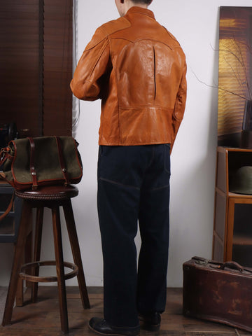 A man wearing a brown leather jacket and dark trousers, photographed from behind in a styled interior setting with a stool, bag, and wooden furniture.