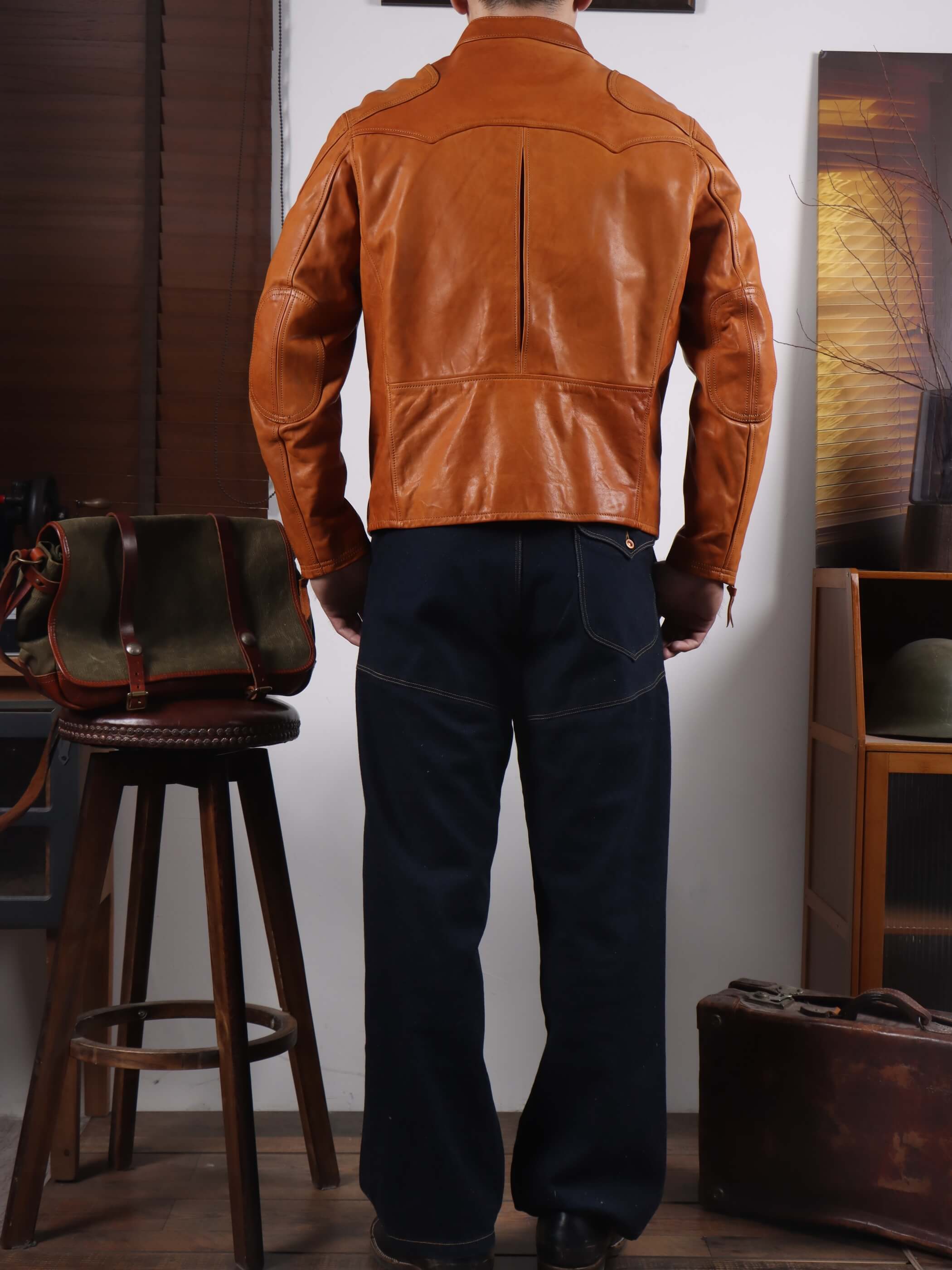A man viewed from behind wearing a tan leather jacket and dark blue jeans, standing in a room with a stool, leather bag, and wooden furniture.
