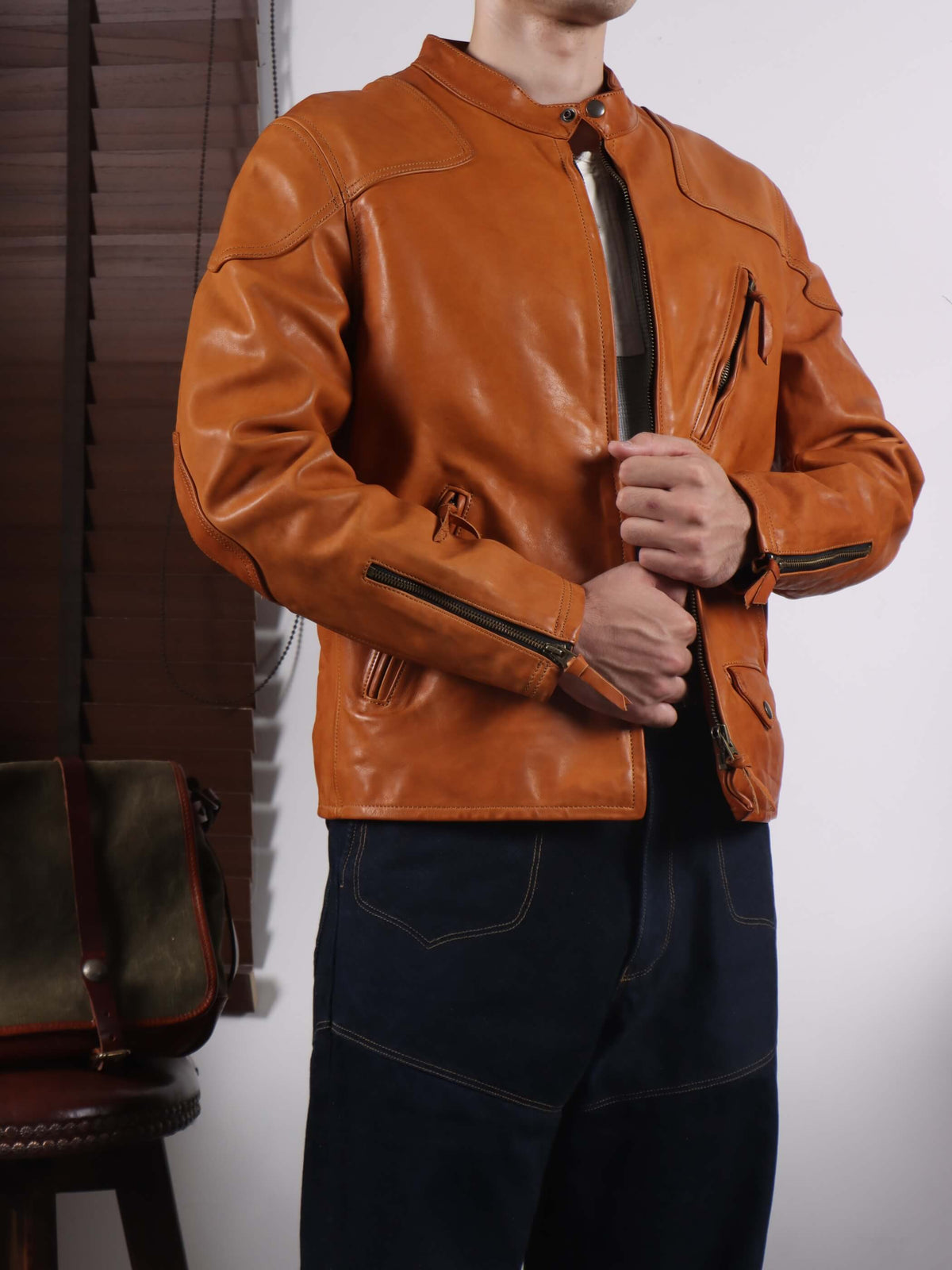 A person wearing a tan-brown leather motorcycle jacket with black zippers, paired with dark blue jeans, standing in front of a white wall.