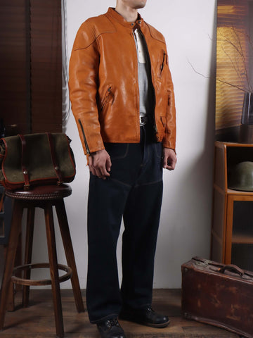 A man wearing a tan leather motorcycle jacket over a white shirt and dark blue jeans, standing in a rustic room with a wooden stool, a vintage suitcase, and a leather bag.