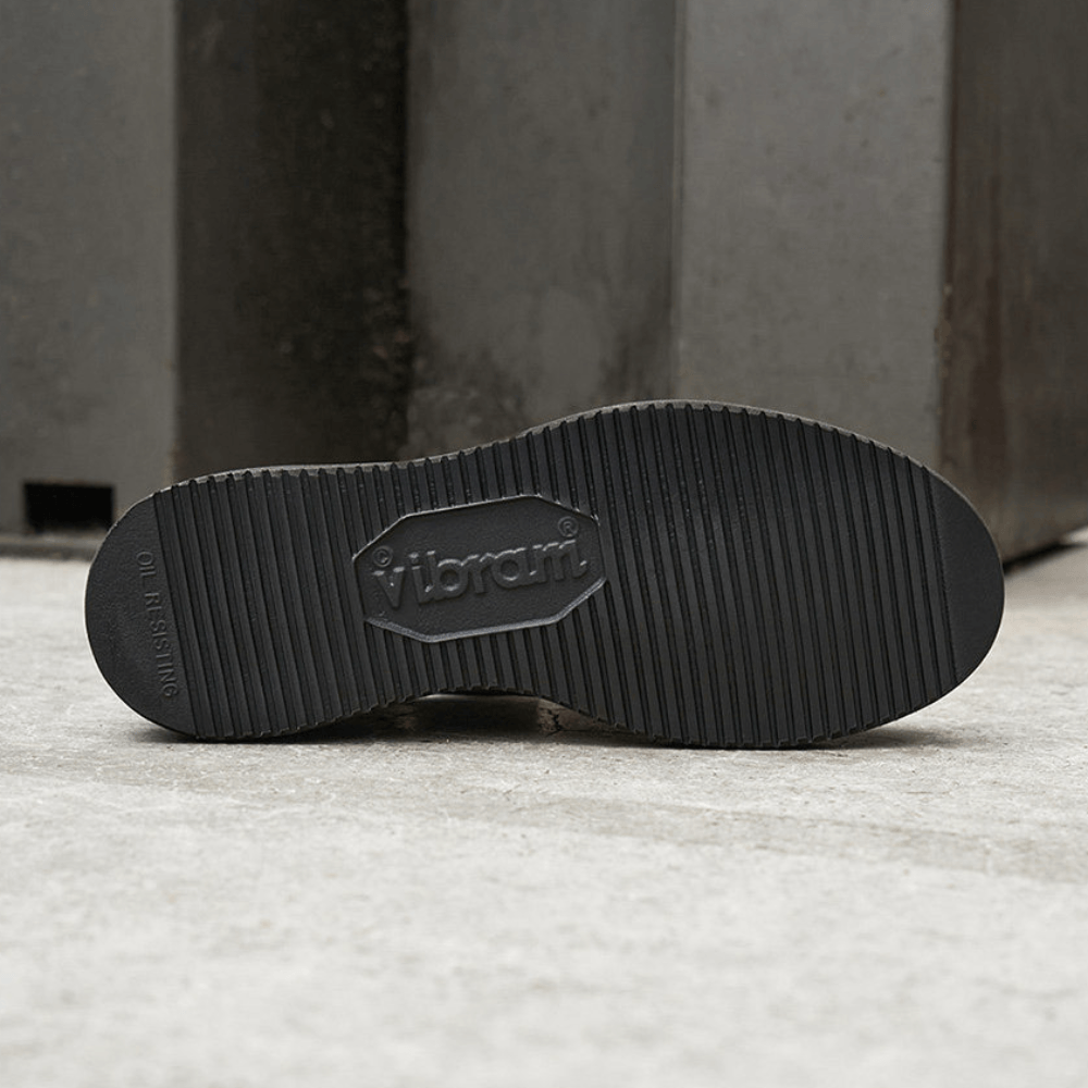 Close-up of the black Vibram outsole of a shoe, showing the embossed Vibram logo and tread pattern, set against a gray concrete background.
