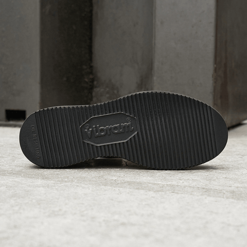 Close-up of the black Vibram outsole of a shoe, showing the embossed Vibram logo and tread pattern, set against a gray concrete background.