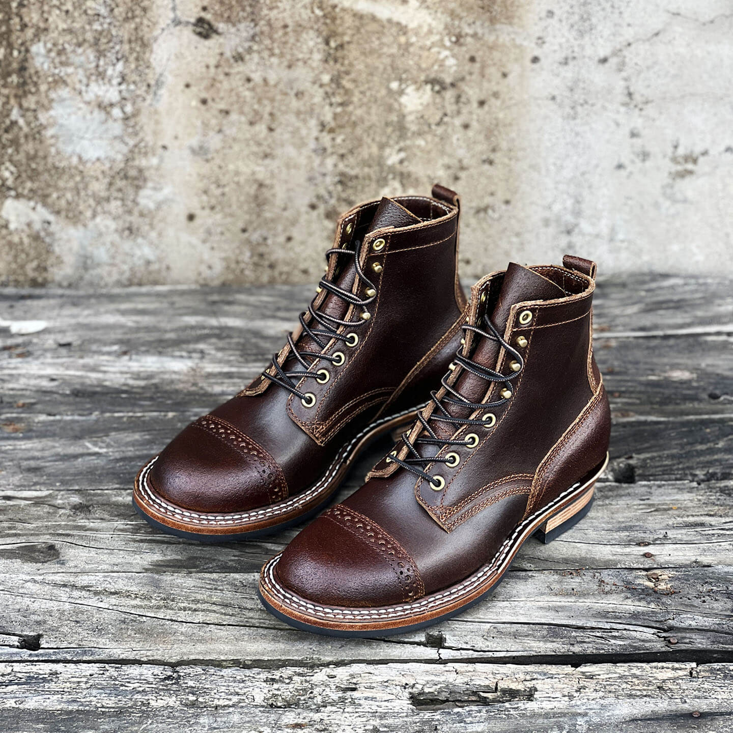 A pair of dark brown leather lace-up boots with cap toes and decorative stitching, displayed on a rustic weathered wooden surface against a textured concrete wall.