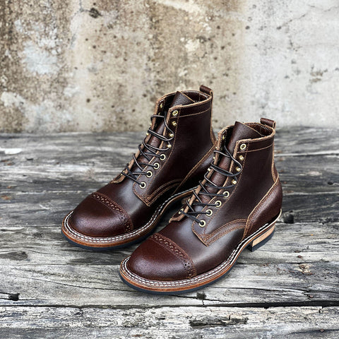 A pair of dark brown leather lace-up boots with cap toes and decorative stitching, displayed on a rustic weathered wooden surface against a textured concrete wall.