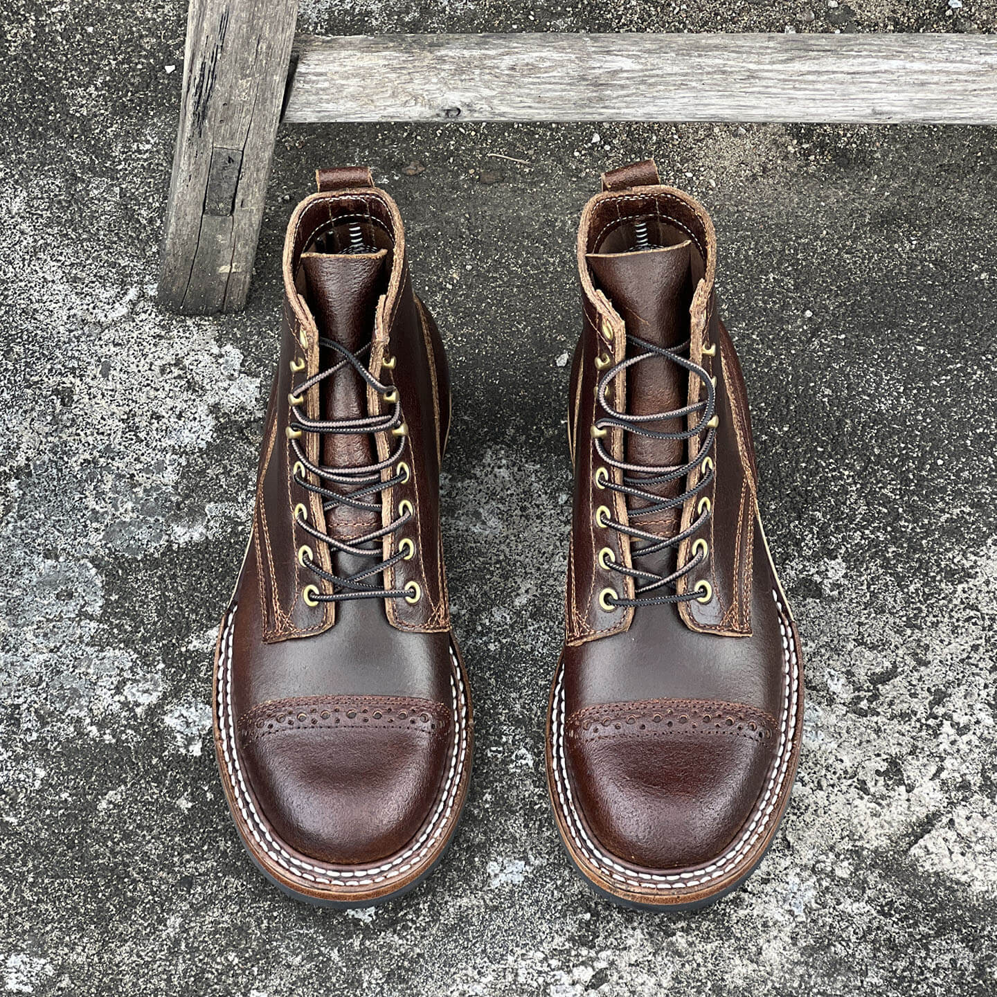 A pair of dark brown leather lace-up boots with brogue detailing and metal eyelets, photographed from above on a textured concrete surface with a weathered wooden post.