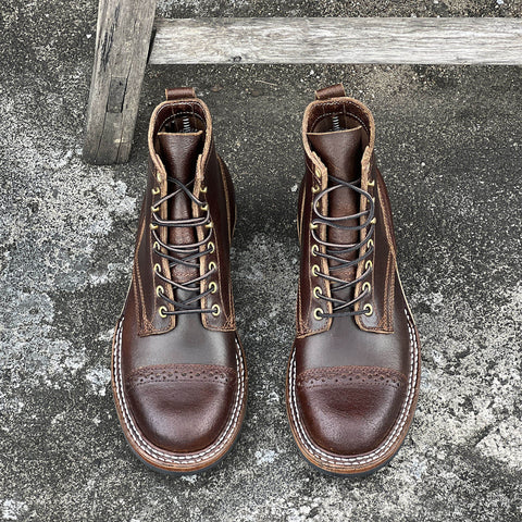 A pair of dark brown leather lace-up boots with brogue detailing and metal eyelets, photographed from above on a textured concrete surface with a weathered wooden post.