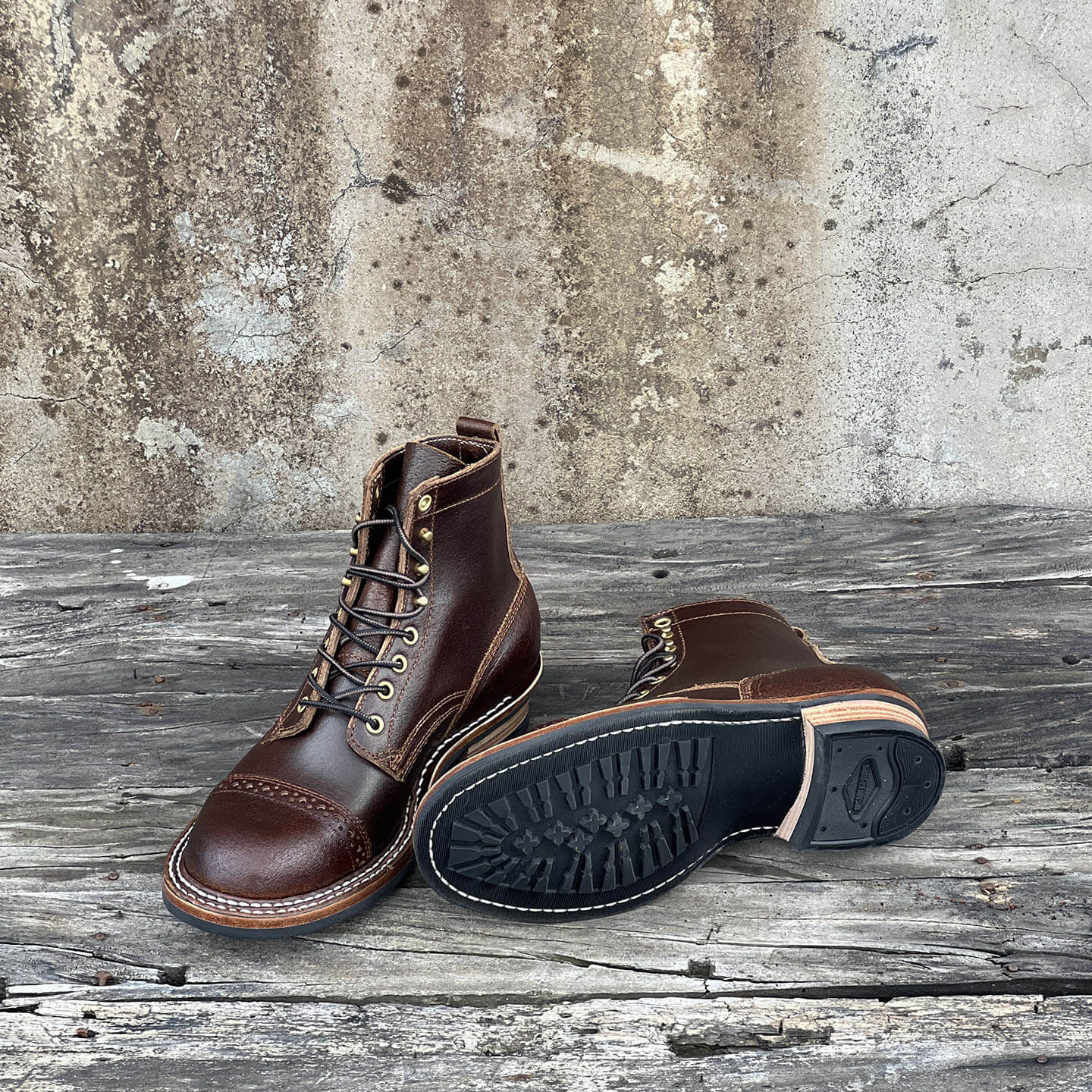 A pair of dark brown leather lace-up work boots, with one boot standing upright showing the side and tongue, and the other lying on its side to display the black rubber sole. They are placed on a weathered wooden plank surface against a textured concrete wall.