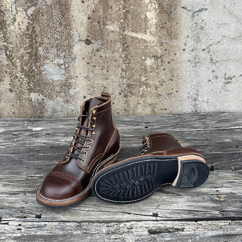 A pair of dark brown leather lace-up work boots, with one boot standing upright showing the side and tongue, and the other lying on its side to display the black rubber sole. They are placed on a weathered wooden plank surface against a textured concrete wall.