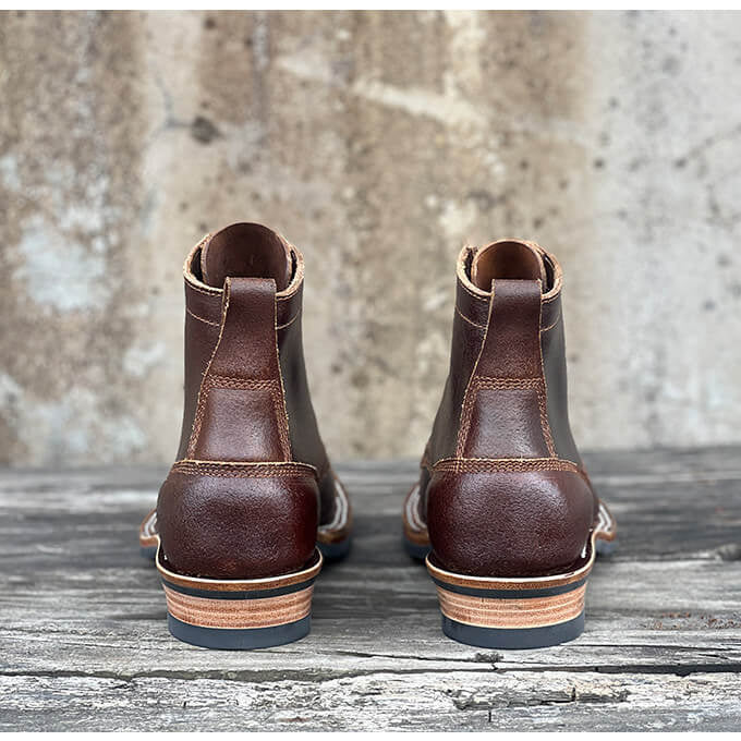 A pair of dark brown leather ankle boots shown from the back, resting on a weathered wooden surface against a textured concrete wall.