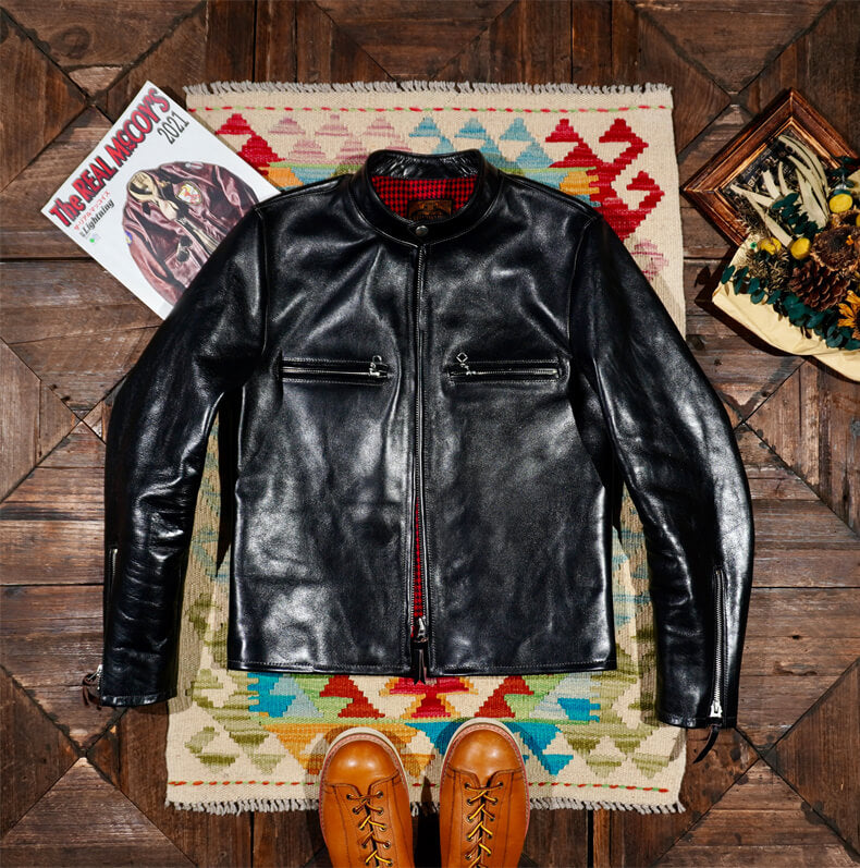 Black leather jacket on a colorful rug with shoes and a magazine on a wooden floor.