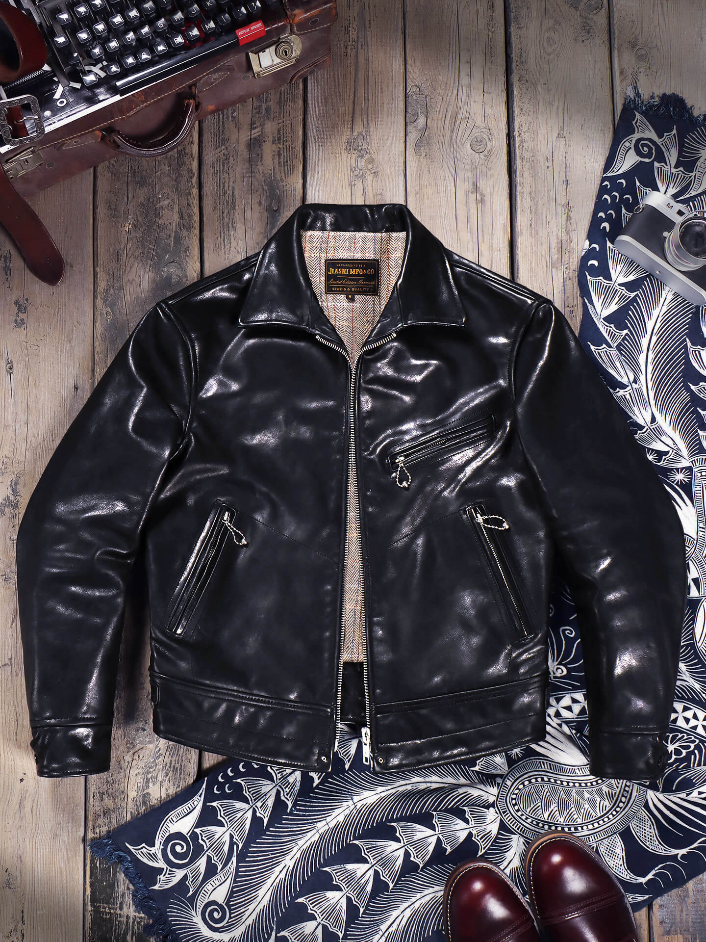 A black leather moto jacket with a high collar and zippered pockets, laid flat on a rustic wooden background with vintage props including a typewriter, patterned fabric, and brown leather shoes.