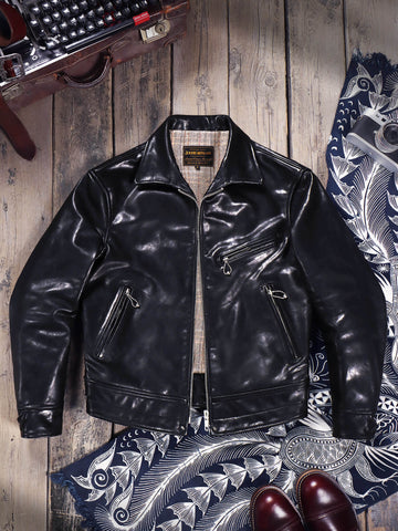 A black leather moto jacket with a high collar and zippered pockets, laid flat on a rustic wooden background with vintage props including a typewriter, patterned fabric, and brown leather shoes.
