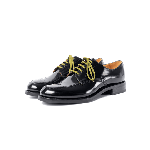 A pair of black polished leather men's derbies with a distinct apron toe design and decorative broad arrows on the vamp. The shoes have yellow laces and a visible goodyear welt.