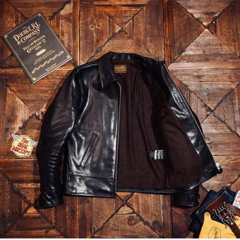 A black leather motorcycle jacket with a brown fleece lining, laid flat on a rustic wooden background with vintage props including a book and a patch.
