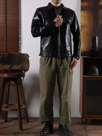 A man wearing a black leather jacket, olive green cargo pants, and black leather shoes, standing in a room with wooden furniture and a stool.