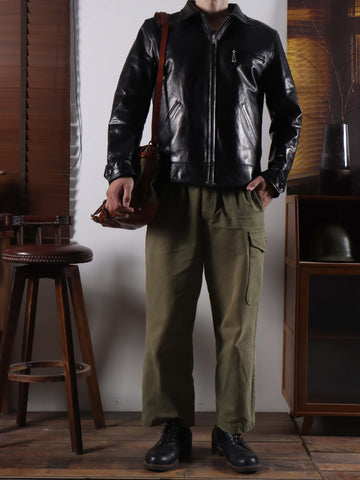 A person wearing a black leather jacket, olive green cargo pants, and black boots, standing in a room with a wooden stool and cabinet. The person is holding a brown leather shoulder bag.